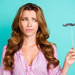 Donna con espressione perplessa tiene un baffo finto, forse riflettendo sulla peluria viso donna.