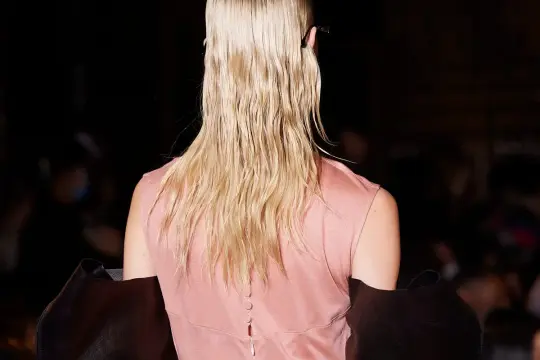 Vista posteriore di una modella con capelli biondi, sottili e pochi, raccolti in una coda bassa. Indossa un abito rosa e un cappotto scuro.