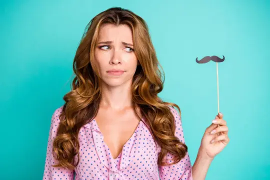 Donna con espressione perplessa tiene un baffo finto, forse riflettendo sulla peluria viso donna.